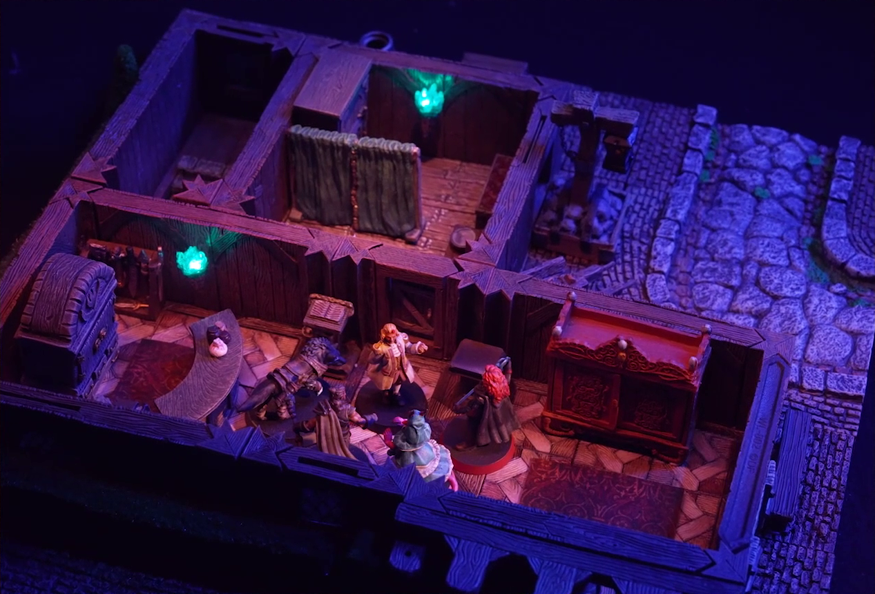 A battlemap of a street-side tailor shop eerily tinted purple and lit by glowing green torches. Kattigan, Wulferic, Tyranny, Bolaire, and Calastro Mercanaud stand in a main entry room in front of a curved front desk. A wardrobe is behind the desk and another one painted red is on the other side of the room. A door leads from the room to an adjoining one with curtains dividing it partially down the center. A third room is off to the side without a clear connection to the rest of the building. This room has stairs leading below ground.