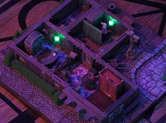 A battlemap of a street-side tailor shop eerily tinted purple and lit by glowing green torches. Kattigan, Wulferic, Tyranny, and Bolaire stand in a main entry room in front of a curved front desk. Calastro Mercanaud hides behind dividing curtains in an adjoining room. A wardrobe is behind the desk and another one painted red is on the other side of the room. A third room is off to the side without a clear connection to the rest of the building. This room has stairs leading below ground. Outside the building is a wheelbarrow.