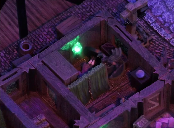 A battlemap of a street-side tailor shop eerily tinted purple and lit by glowing green torches. Kattigan stands over a fallen Calastro Mercanaud behind dividing curtains in one room. Calastro stands just beneath a green torch on the wall. The edge of an adjoining room is visible at the corner of the image. A third room is off to the side without a clear connection to the rest of the building. This room has stairs leading below ground.