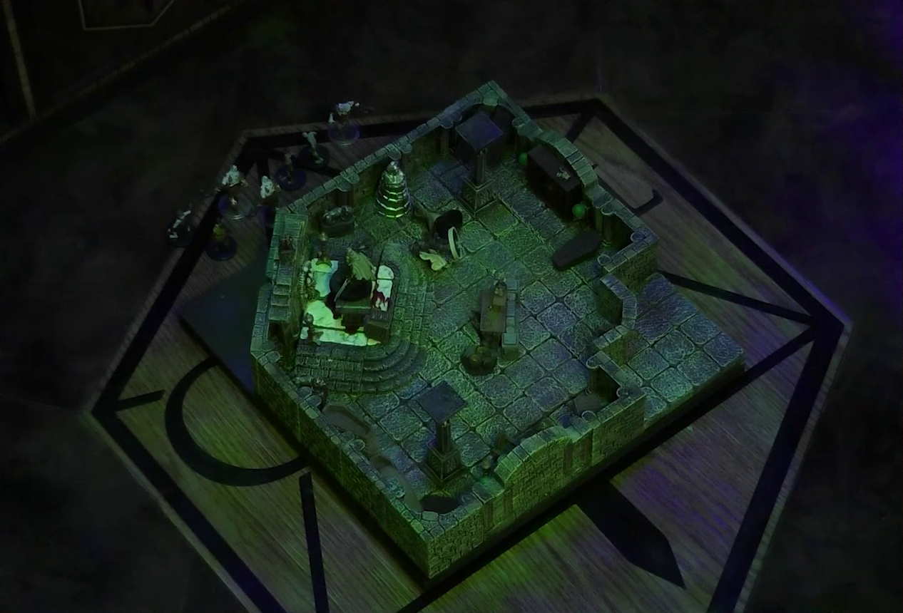 A battlemap of a stone crypt with open wooden coffins on one wall and a silvery metal iron maiden on another wall. Teor and Talcydimir lay downed next to a wooden table on which an undead Raimond Davinos is standing. A hoard of zombies is placed outside the room. The table sits on a raised part of the floor in front of a wall decorated with carved skull motifs. On a box in front of the table sits a tray bearing multiple surgical instruments. The room is lit by eerie glowing green torches.