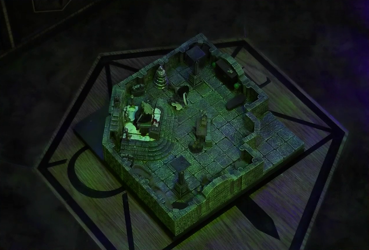 A battlemap of a stone crypt with open wooden coffins on one wall and a silvery metal iron maiden on another wall. Teor and Talcydimir lay downed next to a wooden table on which an undead Raimond Davinos is standing. The hoard of zombies from the previous image is no longer there. The table sits on a raised part of the floor in front of a wall decorated with carved skull motifs. On a box in front of the table sits a tray bearing multiple surgical instruments. The room is lit by eerie glowing green torches.