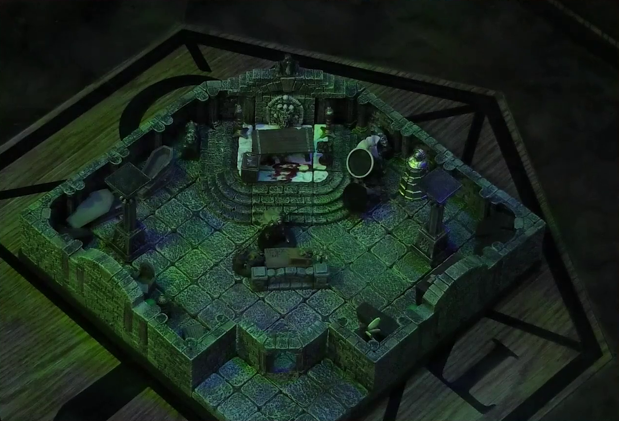 A battlemap of a stone crypt with open wooden coffins on one wall and a silvery metal iron maiden on another wall. An undead Raimond Davinos stands on one side of a short wooden table and stone wall. On the other side, in the corner of the room, is Thimble, off to the side of the open exit to the room. Teor and Talcydimir lay downed next to a wooden table behind Raimond. The table sits on a raised part of the floor in front of a wall decorated with carved skull motifs. The floor under the table is splattered with blood. On a box in front of the table sits a tray bearing multiple surgical instruments. The room is lit by eerie glowing green torches.