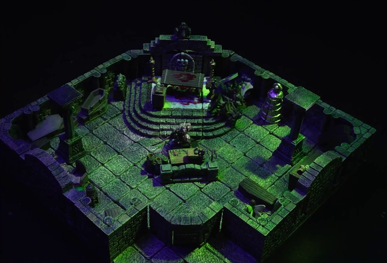 A battlemap of a stone crypt with open wooden coffins on one wall and a silvery metal iron maiden on another wall. An undead Raimond Davinos stands on one side of a short wooden table and stone wall. On the other side, in the corner of the room, is Thimble, off to the side of the closed exit to the room. Teor and Talcydimir lay downed next to a wooden table behind Raimond. The table sits on a raised part of the floor in front of a wall decorated with carved skull motifs. The floor under the table is splattered with blood. On a box in front of the table sits a tray bearing multiple surgical instruments. The room is lit by eerie glowing green torches.