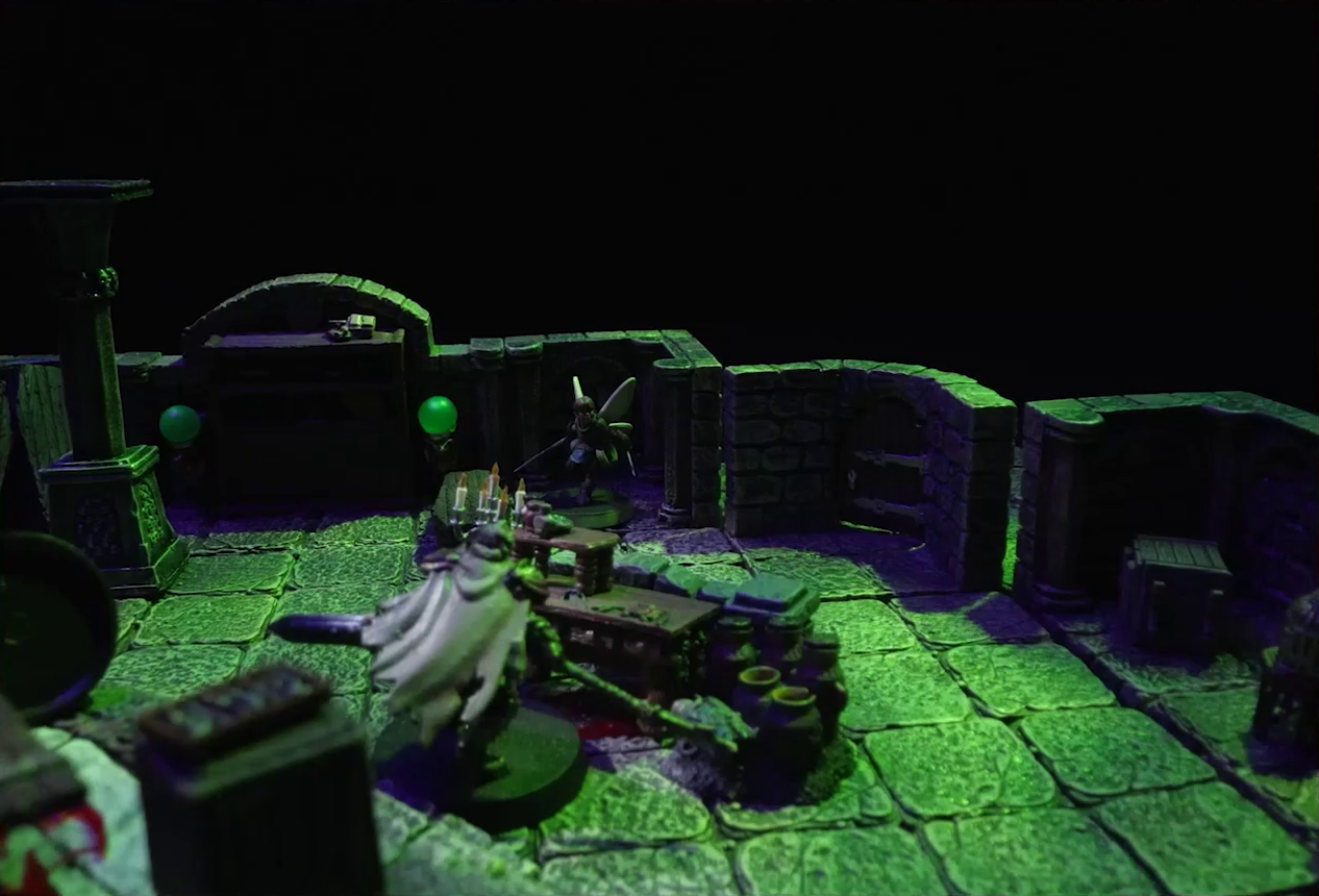 A battlemap of a stone crypt focused on an undead Raimond Davinos standing on one side of a short wooden table and stone wall. On the other side, in the corner of the room, is Thimble, off to the side of the closed exit to the room. At the base of the stone wall are some small pots. On the wooden table is a lit candelabra with five candles. The room is lit by eerie glowing green torches.