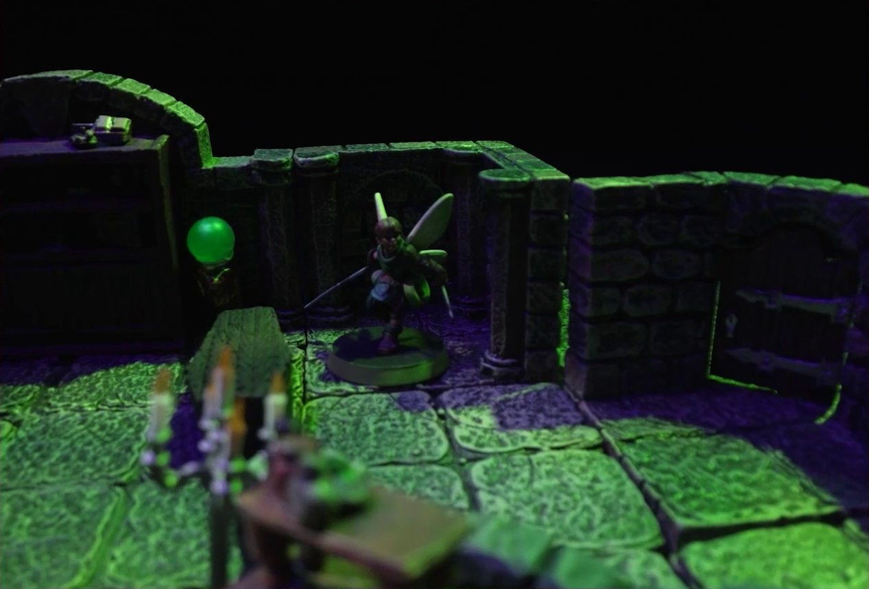 A battlemap of a stone crypt focused on Thimble standing in the corner of the room. Blurry in the foreground is a candelabra with five candles sitting on a wooden table next to a short stone wall. Thimble stands next to a closed wooden coffin and off to the side of a closed wooden door in the wall, exiting the room. The room is lit by eerie glowing green torches.