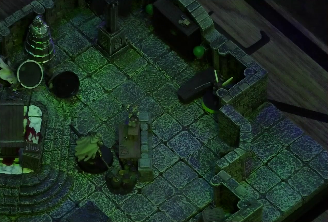 A battlemap of a stone crypt with open wooden coffins on one wall and a silvery metal iron maiden on another wall. An undead Raimond Davinos stands on one side of a short wooden table and stone wall. On the other side, in the corner of the room, is Thimble. Teor and Talcydimir lay downed next to a wooden table behind Raimond. The table sits on a raised part of the floor. The floor under the table is splattered with blood. On a box in front of the table sits a tray bearing multiple surgical instruments. The room is lit by eerie glowing green torches.