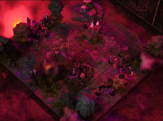 A battlemap of a forest clearing with a large leafless Hawthorn tree near the center. Fake smoke rises from that tree and others at the edges of the map. Orange lights indicate that the trees are on fire. A number of Candescent Creed members and fairy poachers are scattered around the area, especially at the base of the Hawthorn tree. A small animal hide hut is at the edge of the battlemap. Large red mushrooms with white spots and a huge purple flower are at the other end near a lake.