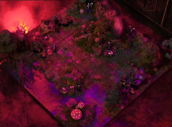 A battlemap of a forest clearing with a large leafless Hawthorn tree near the center. Fake smoke rises from that tree and others at the edges of the map. Orange lights indicate that the trees are on fire. A number of Candescent Creed members and fairy poachers are scattered around the area, especially at the base of the Hawthorn tree. A small animal hide hut is at the edge of the battlemap. Large red mushrooms with white spots and a huge purple flower are at the other end near a lake.