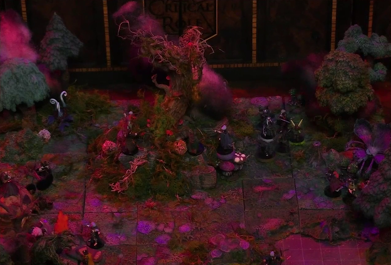 A battlemap of a forest clearing with a large leafless Hawthorn tree near the center. Fake smoke rises from that tree and others at the edges of the map. Thimble, Kattigan, Wulferic, Vaelus, Occtis, Pin, and Ulbid Morn stand near the tree. A number of Candescent Creed members and fairy poachers are scattered around the area, especially at the base of the Hawthorn tree. A huge purple flower is at the edge of the map near a lake.