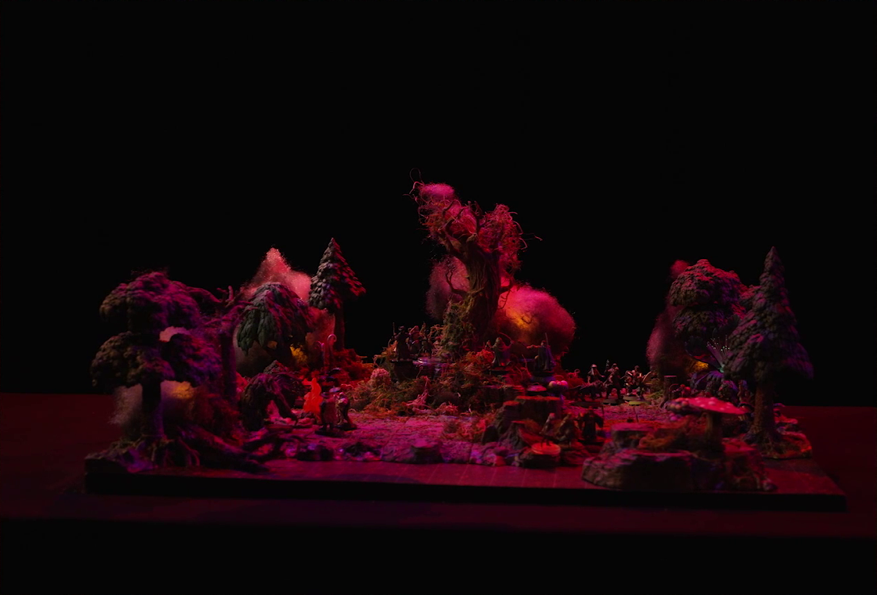 A battlemap of a forest clearing with a large leafless Hawthorn tree near the center, bathed in an eerie red glow. Fake smoke rises from that tree and others at the edges of the map. Orange lights indicate that the trees are on fire. Thimble, Kattigan, Wulferic, Vaelus, Occtis, Pin, and Ulbid Morn stand near the tree. A number of Candescent Creed members and fairy poachers are scattered around the area, especially at the base of the Hawthorn tree. Large red mushrooms with white spots and a huge purple flower are at the edge of the battlemap near a lake.