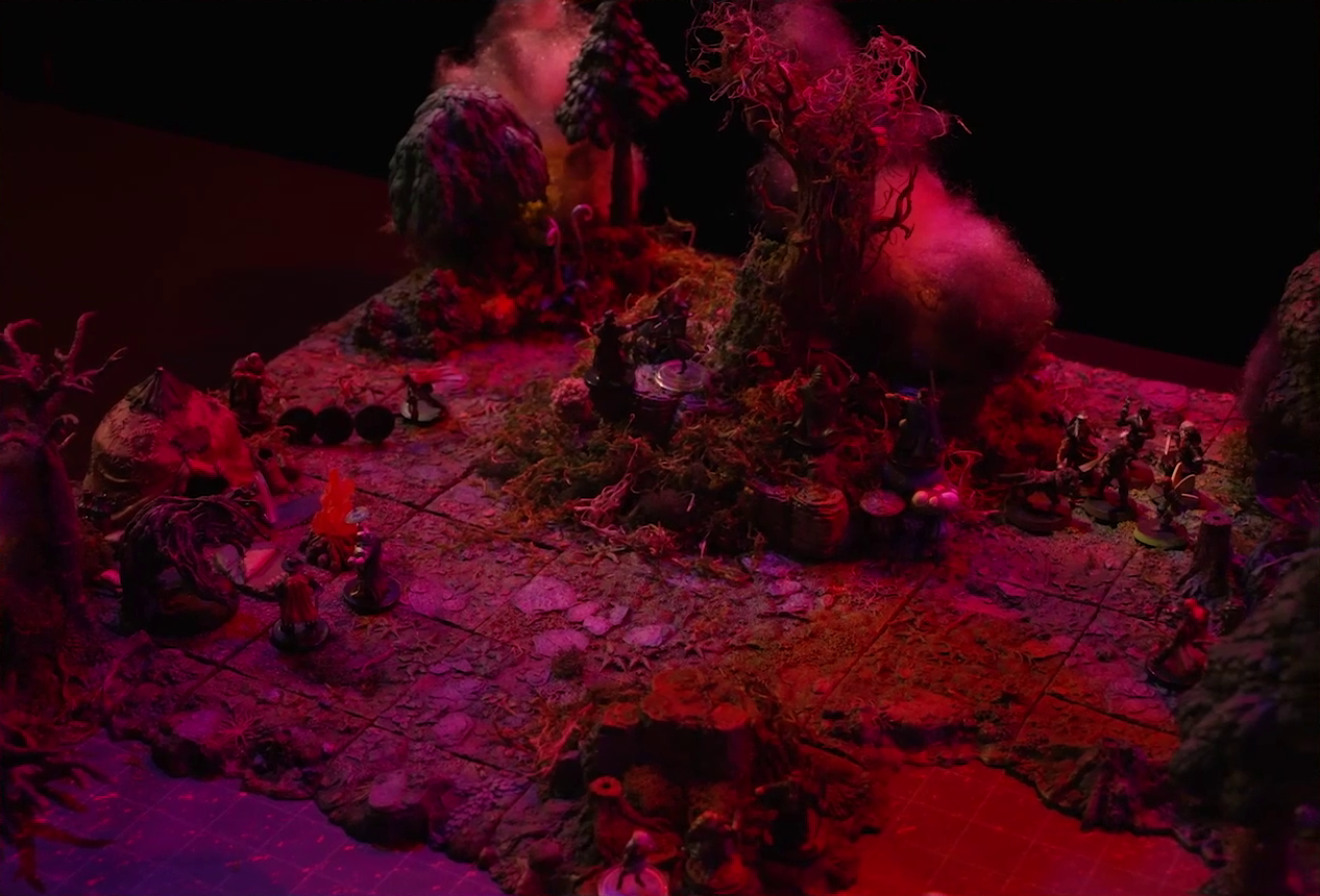 A battlemap of a forest clearing with a large leafless Hawthorn tree near the center, bathed in an eerie red glow. Fake smoke rises from that tree and others at the edges of the map. Orange lights indicate that the trees are on fire. Thimble, Kattigan, Wulferic, Vaelus, Occtis, Pin, and Ulbid Morn stand near the tree. A number of Candescent Creed members and fairy poachers are scattered around the area, especially at the base of the Hawthorn tree. A small animal hide hut is at the edge of the battlemap. Large red mushrooms with white spots and a huge purple flower are at the other end near a lake.