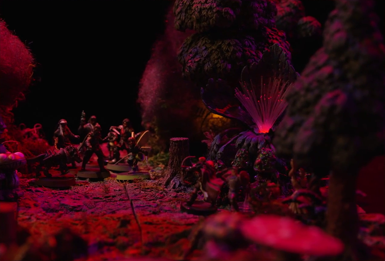 A battlemap of a forest clearing bathed in an eerie red glow. Fake smoke rises from the trees at the edges of the map. Orange lights indicate that the trees are on fire. Thimble, Kattigan, Wulferic, Vaelus, Occtis, Pin, and Ulbid Morn stand together and a number of Candescent Creed members and fairy poachers are scattered around the area. A huge purple flower with long fibers emerging from it emits a red glow.