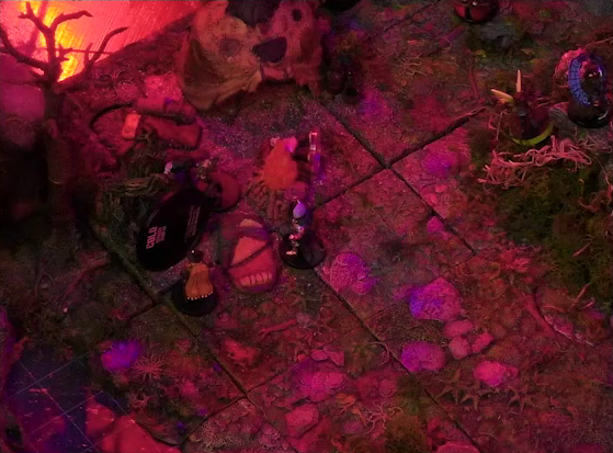 A battlemap of a forest clearing with a leafless tree at the edge bathed in an eerie red glow. Fake smoke rises from the tree and orange lights indicate it’s on fire. The shambling mound Shuffa lies at the foot of the tree, surrounded by Candescent Creed members and fairy poachers. A small animal hide hut is nearby. Thimble faces off against a Candescent Creed member with a “DEAFENED” status ring around her miniature at the base of a large tree at the edge of the image.