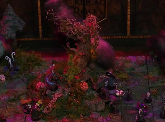 A battlemap of a forest clearing focused on a large leafless Hawthorn tree. Fake smoke rises from that tree and others at the edges of the map. Orange lights indicate that the trees are on fire. Thimble, Kattigan, Wulferic, and Gaya Seremai face off against Candescent Creed members and fairy poachers at the base of the Hawthorn tree. One Candescent Creed member has a “DEAFENED” status ring around her miniature. Ulbid Morn and Vaelus stand a short distance away.