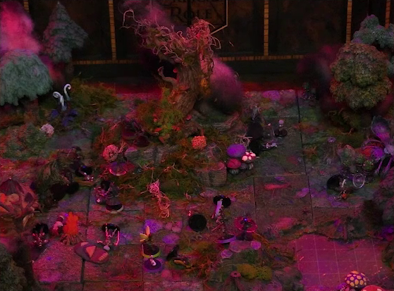 A battlemap of a forest clearing with a large leafless Hawthorn tree near the center. Fake smoke rises from that tree and others at the edges of the map. Orange lights indicate that the trees are on fire. A number of Candescent Creed members and fairy poachers are scattered around the area, most downed. Thimble, Kattigan, Wulferic, Occtis, Pin, Vaelus, Julien, and Ulbid Morn are likewise scattered around, having completed their fight. Large red mushrooms with white spots and a huge purple flower are at one edge of the battlemap near a lake.