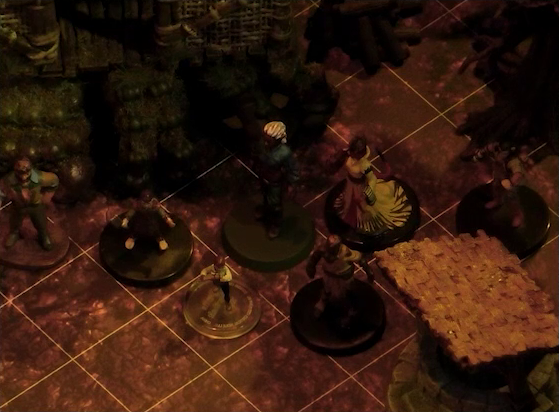 A battlemap focused on Rei’nia standing in a group of six villagers. They stand next to a covered stone well and a wooden building on a rocky platform.
