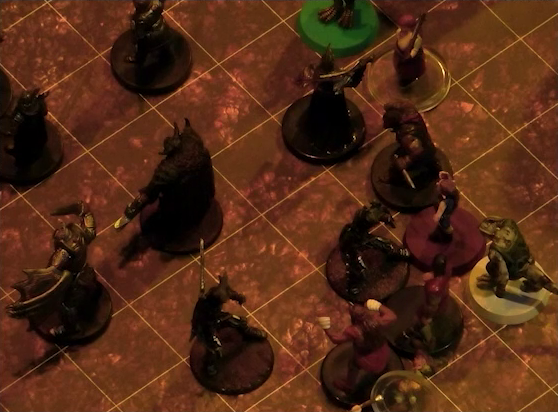 A battlemap focused on a group of six black-armored soldiers surrounding a commander wearing a horned helmet. Crokas, Fiedra, Erro, and a number of villagers face off against the soldiers. One villager lays on the ground.
