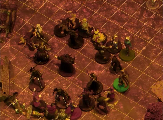 A battlemap focused on a group of six black-armored soldiers surrounding a commander wearing a horned helmet. Surrounding the soldiers are Garen, Rei’nia, Erro, Fiedra, Crokas, and a number of villagers. Three villagers lay on the ground near the soldiers. Some wooden debris and trees in autumn colors are visible at the edge of the image.
