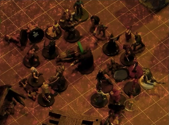 A battlemap focused on a fight between a group of black-armored soldiers and Garen, Rei’nia, Fiedra, Crokas, and a number of villagers. Erro lays on the ground in the center of the fight, with a number of villagers also downed. A soldier lays on the ground in front of Fiedra and Crokas. Some wooden debris is visible at the edge of the image.