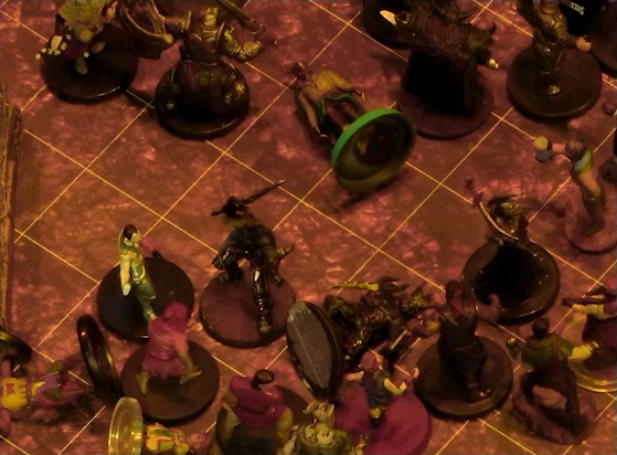 A battlemap focused on a fight between a group of black-armored soldiers and Fiedra, Crokas, and a number of villagers. Erro lays on the ground in the center of the fight, with a number of villagers also downed. A soldier lays on the ground in front of Fiedra and Crokas.