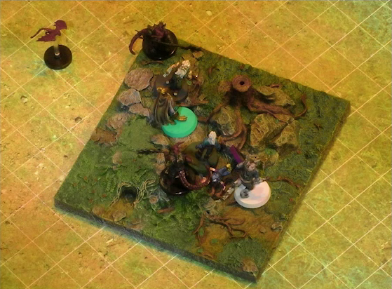 A battlemap of a grassy plain focused on a textured map square, on which are Garen, Erro, Rei’nia, Fiedra, and Crokas facing off against two dragon-like devils and a red flying impish creature. Fiedra is fallen next to one devil. The devils are armed with barbed pole weapons. The map square is mostly covered in green, with some rocks and a single tree stump.