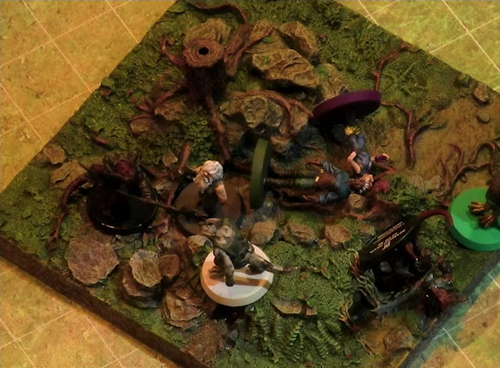 A battlemap of a grassy plain focused on a textured map square, on which are Garen, Erro, Rei’nia, Fiedra, and Crokas facing off against two dragon-like devils. Fiedra and Rei’nia are fallen, as is one devil. The devils are armed with barbed pole weapons. The map square is mostly covered in green, with some rocks and a single tree stump.