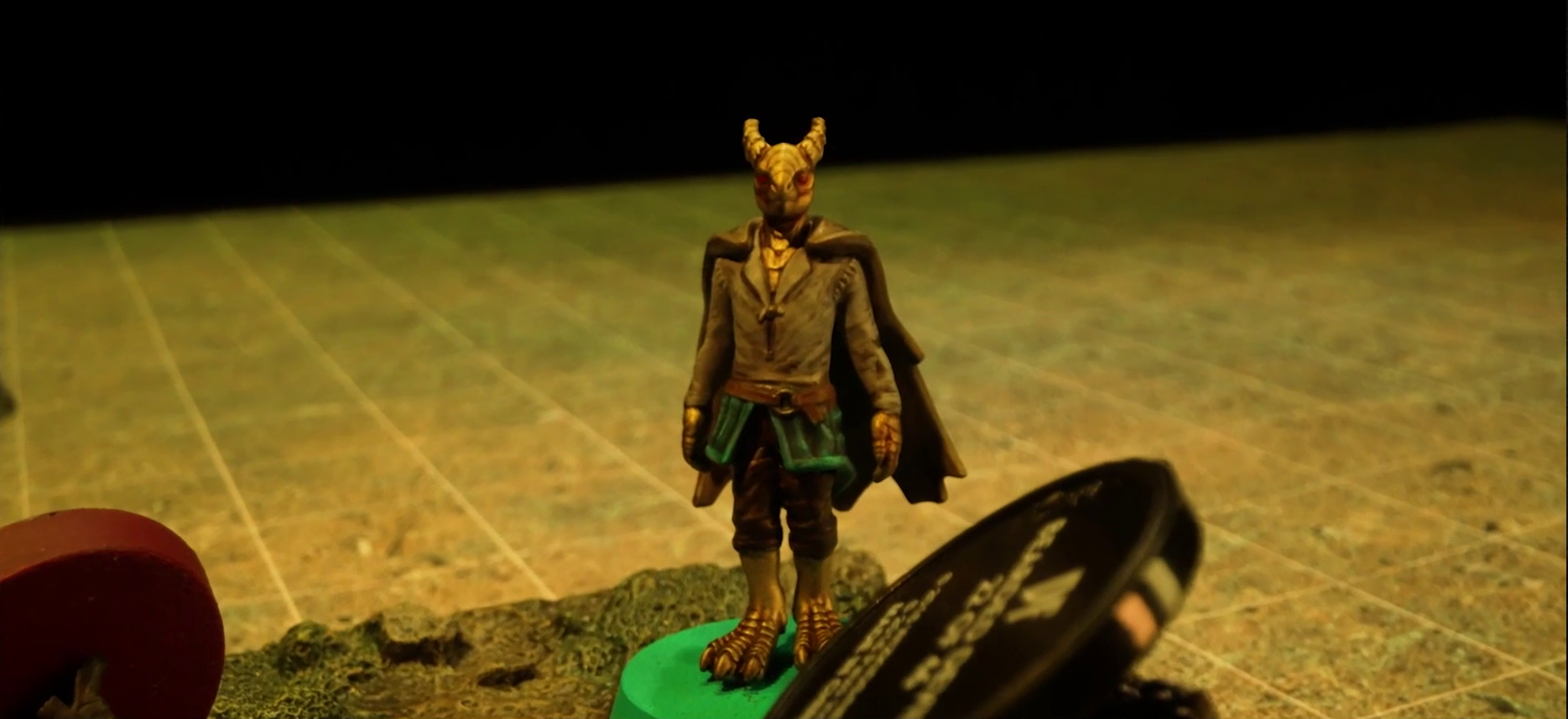 A battlemap focused on Erro standing on a textured map square in the middle of a grassy plain. Before him is the base of Fiedra’s miniature, laying on the ground, and the black base of another miniature also fallen.