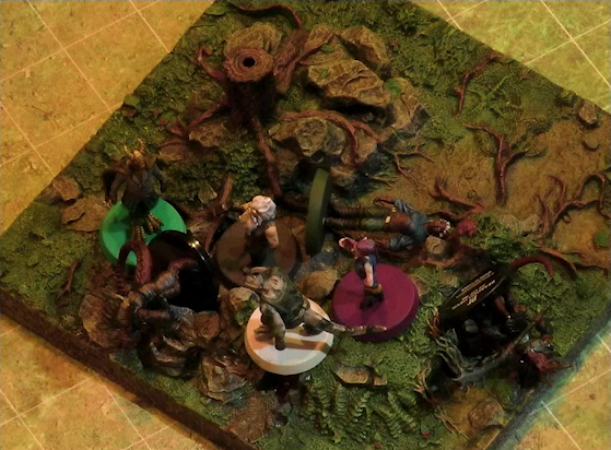 A battlemap of a grassy plain focused on a textured map square, on which are Garen, Erro, Rei’nia, Fiedra, and Crokas standing next to two fallen dragon-like devils. Rei’nia is also fallen. The devils are armed with barbed pole weapons. The map square is mostly covered in green, with some rocks and a single tree stump.