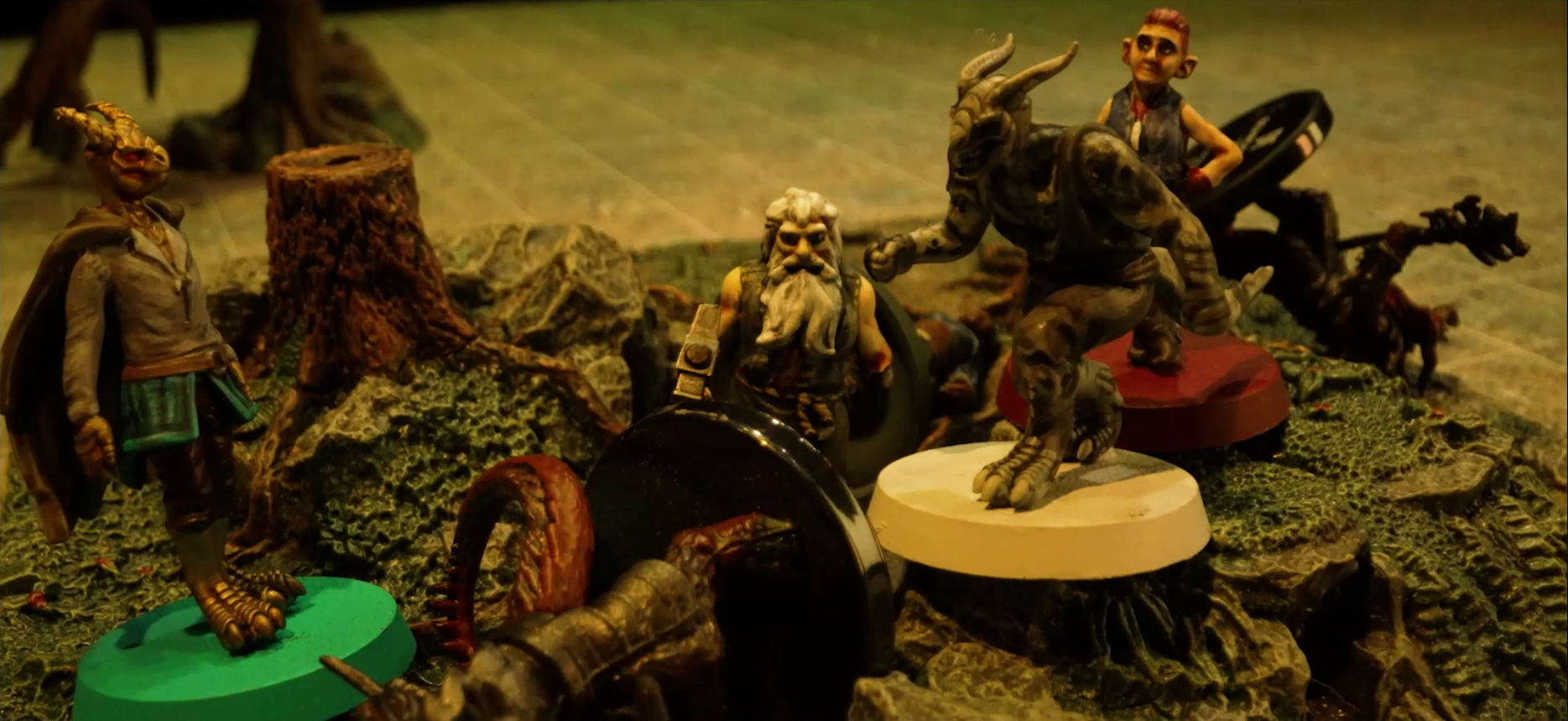 A battlemap focused on Erro, Garen, Crokas, and Fiedra standing on a textured map square in the middle of a grassy plain. Rei’nia is fallen next to Fiedra, as are two dragon-like devils armed with barbed pole weapons. The map square is mostly covered in green, with some rocks and a single tree stump. A single tree is visible at the edge of the image in the otherwise empty plain.
