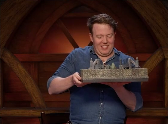 Brennan carefully carrying a battlemap of a walled stone chamber to the Critical Role table. A number of pillars and arches peek over the top of the walls. Brennan is smiling in nervous concentration, and he is wearing a blue denim button-up.