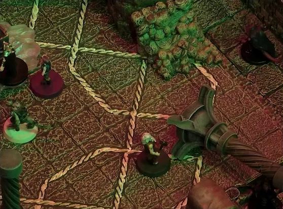 A battlemap of a stone temple interior focused on a toppled stone pillar, next to which is Garen. Crokas, Fiedra, and Lord Callister are just visible at the left edge of the image. A red tailed devil is a short distance away. Garen wields a metal hammer in his bare muscular right arm and sports a white beard and hair.