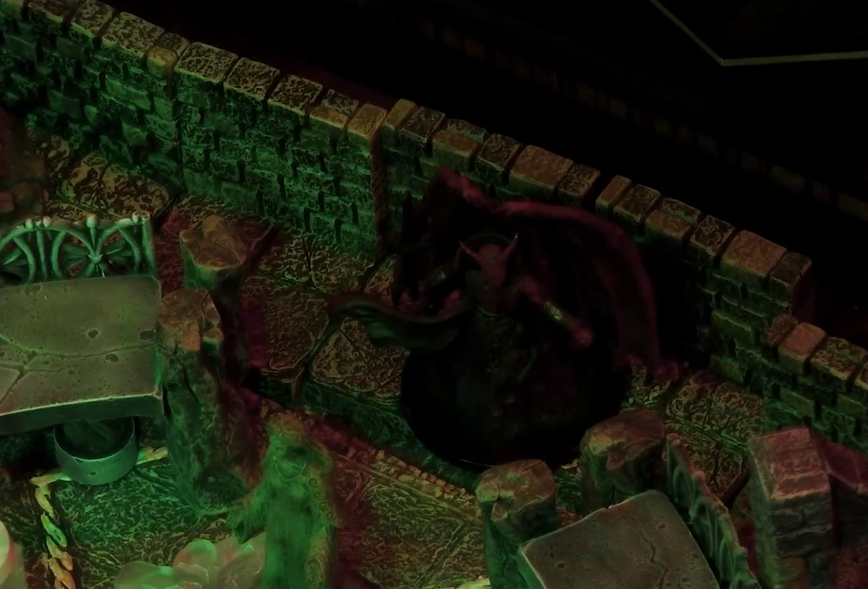 A battlemap of a stone temple interior focused on a miniature of Asmodeus. He stands against the wall behind a statue of the Moonweaver. He has red skin, horns, and huge wings which unfurl behind him. He wields a trident. A cape flows in front of him from behind his back.
