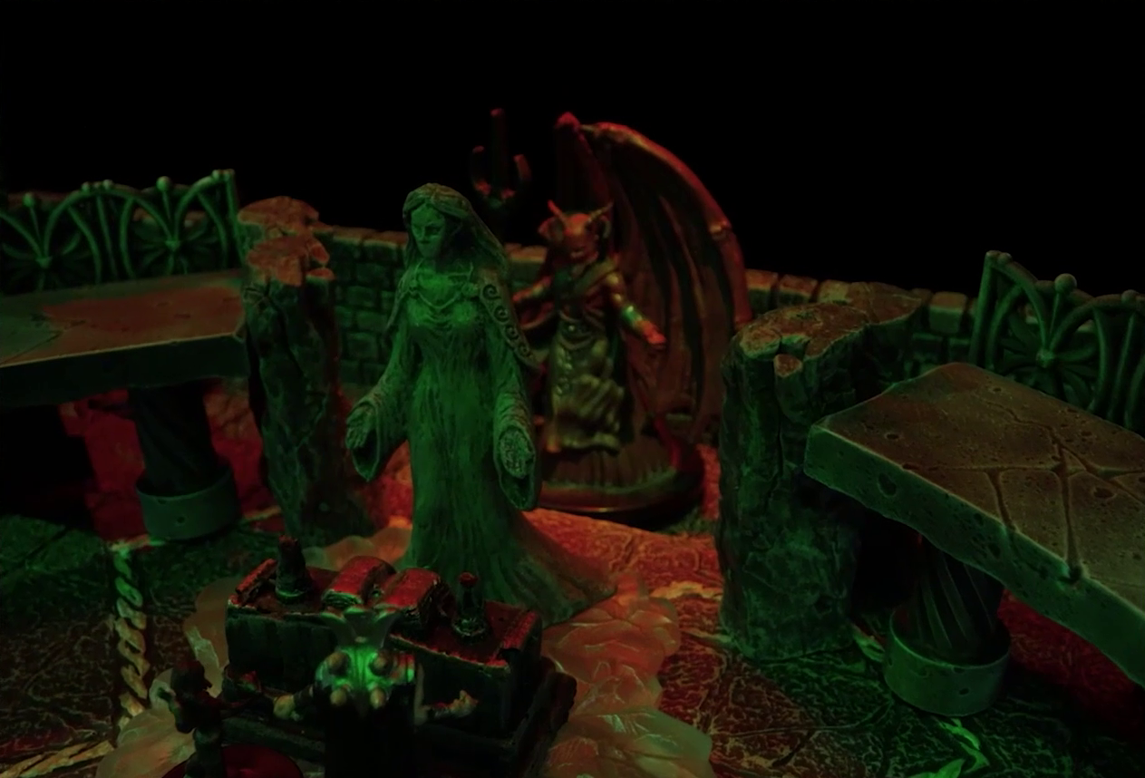 A battlemap of a stone temple interior focused on a statue of the Moonweaver, behind which is Asmodeus. Asmodeus has red skin, horns, and huge wings which unfurl behind him. He wields a trident and wears a flowing gray robe. In front of the statue is an altar with a book open on it. Timothy and Lord Callister stand before the altar.
