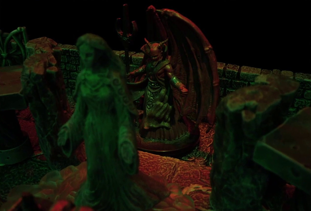 A battlemap of a stone temple interior focused on a statue of the Moonweaver, behind which is Asmodeus. Asmodeus has red skin, horns, and huge wings which unfurl behind him. He wields a trident and wears a flowing gray robe.