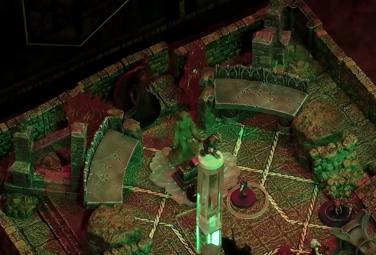 A battlemap of a stone temple interior. Admodeus stands, huge wings behind him, at the rear of the room behind a statue of the Moonweaver. On either side of him are massive fires. Crokas levitates in the air. On the ground are Timothy, Lord Callister, Fiedra with a status ring, Garen, and a red devil with metallic spikes and pincers. Garen stands in front of a toppled stone pillar.