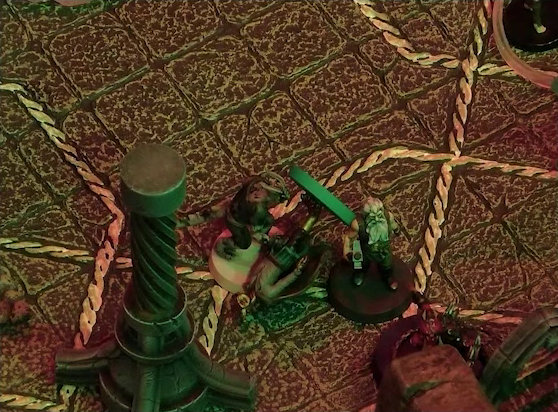 A battlemap of a stone temple interior focused on Crokas and Garen standing to either side of a fallen Erro. Garen faces a red devil with metallic spikes and pincers. Next to them is a stone pillar with a helical pattern of lines running up its length. Just visible at the top edge of the image is another devil.