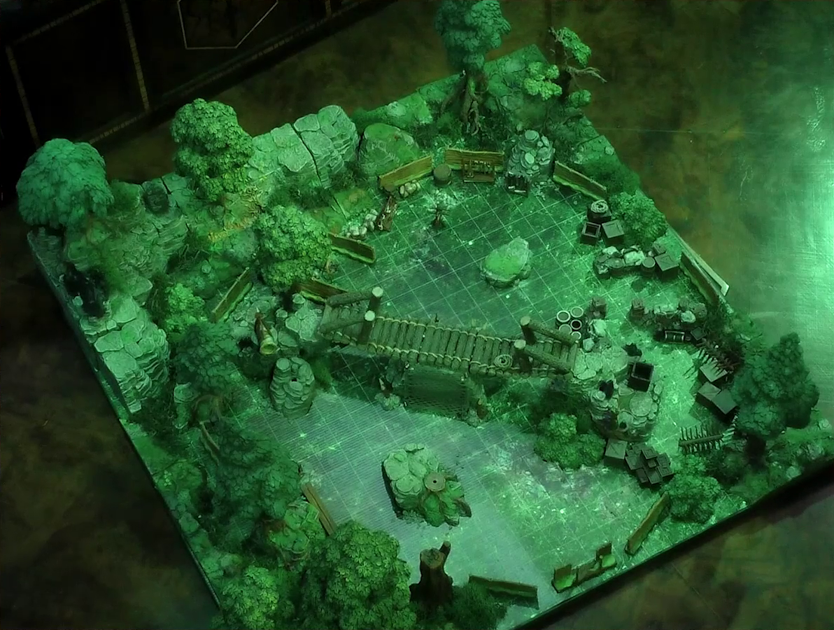 A battlemap of a forest clearing cut down the center with a log bridge spanning two stone ledges. Rocky outcroppings border two sides of the map. A number of boxes, crates, and wooden barriers are strewn throughout.