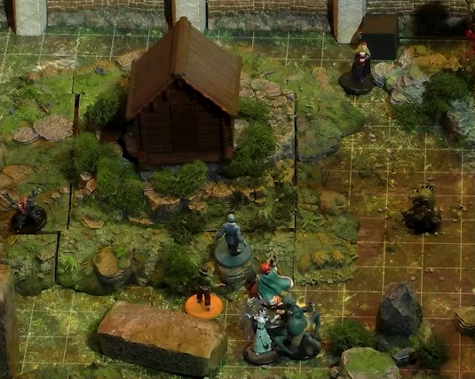 A close-up of a battlemap showing a section of a wooded courtyard with a stone defensive wall at the top edge of the image. A small wooden shed sits among some bushes in the center of the courtyard. Kai, Jessep, and Sky approach the shed. Behind and around them are five creatures. A small ball-shaped creature with its torso split open in a toothy maw, two clawed hands emerging from its headless body. A robed humanoid surrounded by thick translucent strands. A creature with teal skin and four spider-like legs propping up a torso with two arms and a rodent-like head. A small reptilian creature standing on two clawed feet with spines running down its back. And a human-looking individual wearing a cloak.