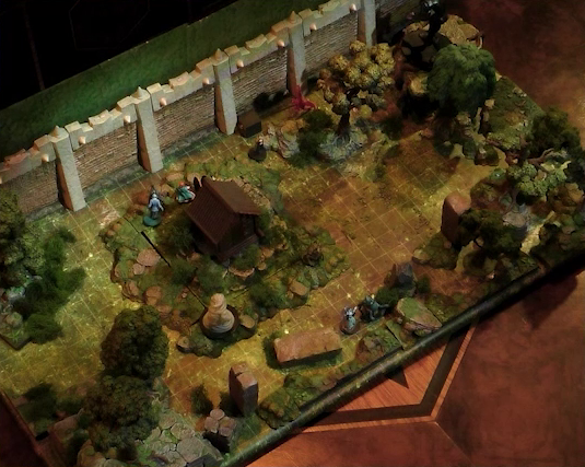 A battlemap of a wooded courtyard bordered on one side by a tall stone defensive wall. A translucent blue magic-looking barrier tops the wall. A small wooden shed sits among some bushes in the center of the courtyard. Filling the rest of the space are trees and old-looking standing stones with circular runic patterns carved into their sides. Jessep and Sky are visible behind the shed. A number of other miniatures are spread throughout, including a flying red creature and a pillar of swirling wind.
