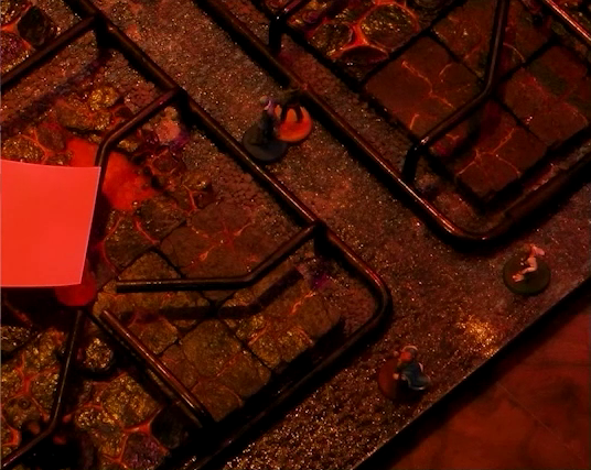 A close-up of a battlemap that looks like a stovetop. Jessep and Kai stand between two burners. The one on the left has a pink sticky note over the center. Sky and Padmund stand at the edge of the stove. The base of the battlemap looks like magma and the scene is bathed in red light.