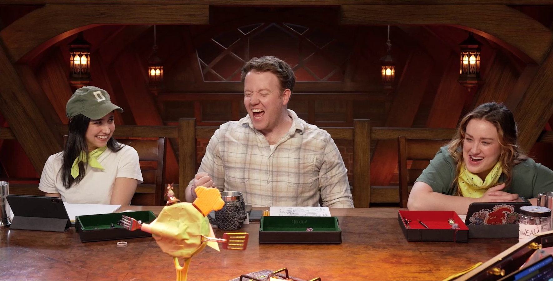 Libe Barer, Brennan Lee Mulligan, and Ashley at the table wearing their yellow Wildemount Wildlings bandanas. Libe is smiling at Brennan, Brennan is cheering and gesturing with a fist, and Ashley is looking forward smiling. In the foreground is visible the back of Wok Wok, the giant frying pan creature. A miniature of Beau stands on its head.