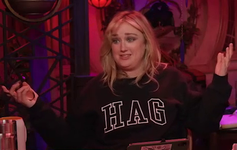 Ashley sitting at the table wearing a black sweatshirt with the text “HAG” in large white collegiate font across the front. Ashley shrugs in such a way as to draw attention to her sweatshirt. The set is tinted a foreboding red as if lit by a fiery glow.