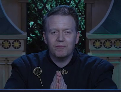 Brennan is wearing a black formal shirt with a gold pin of a hand with the index finger extended, dressed as Tywin Lannister from Game of Thrones. He sits at the table behind the DM screen, his hands pressed together as he stares forward menacingly.