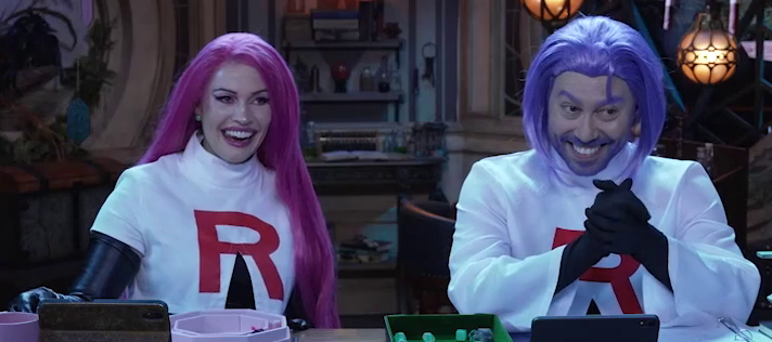 Whitney and Sam at the table dressed in Halloween costumes. They wear pink and blue wigs and white crop tops with red “R”s on them, dressed as Jessie and James from Team Rocket. Both have long black gloves on their hands. Whitney is laughing, Sam clasps his hands in front of him as if scheming.