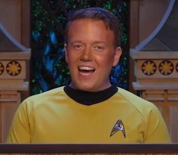 Brennan wearing a yellow long-sleeve shirt, dressed as Captain Kirk from Star Trek. He sits at the table behind the DM screen and grins heroically.