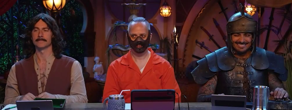 Matt, Taliesin, and Robbie at the table dressed in Halloween costumes. Matt is in a vest, long hair, and moustache, dressed as Inigo Montoya from the Princess Bride. Taliesin wears an orange jumpsuit and metal muzzle, dressed as Hannibal Lecter from The Silence of the Lambs. Robbie is in a suit of armor and feather plume helmet, dressed as Odysseus from the Odyssey.
