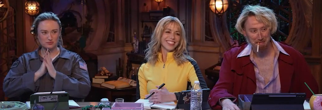 Ashley, Laura, and Alexander at the table dressed in Halloween costumes. Ashley is in an astronaut outfit with a headset, dressed as Ellie Arroway from Contact. Laura is in a yellow and black jumpsuit, dressed as Beatrix Kiddo from Kill Bill. Alexander wears a red trenchcoat, smokes a cigarette, and has scars across his face, dressed as Masao Kakihara from Ichi the Killer.