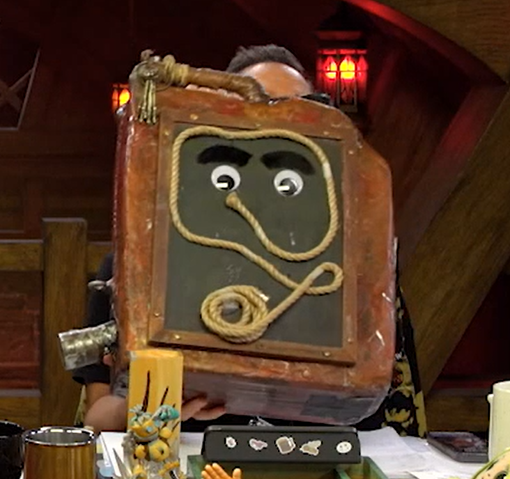 A rusty gas can with a thick rope pasted to it. The rope is coiled at the bottom and formed into a square shape at the top. Two large googly eyes with bushy eyebrows are pasted within the square.