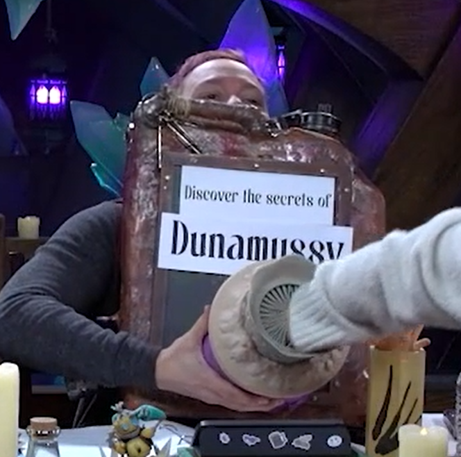 A rusty gas can with “Discover the secrets of Dunamussy” on white paper at the top. Sticking out from the can below is a purple tube with a tapered top leading to an opening with many teeth, like a sand worm. Sam is supporting the tube from below and Laura’s hand is reaching into the mouth.