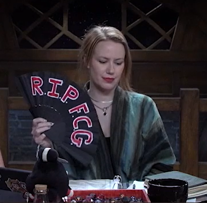 Marisha holding a black hand fan
            with red text outlined in white reading “R.I.P. F.C.G.” She is
            looking down sadly.
